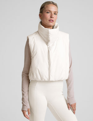 model wearing a white puffer vest over a long-sleeve shirt on a light gray background. 