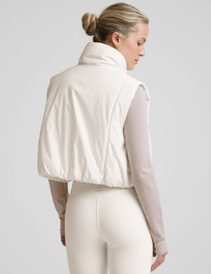 model wearing a white puffer vest over a long-sleeve shirt on a light gray background. 