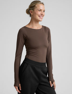 woman wearing a brown long-sleeve boat neck bodysuit and black pants against a plain background. 