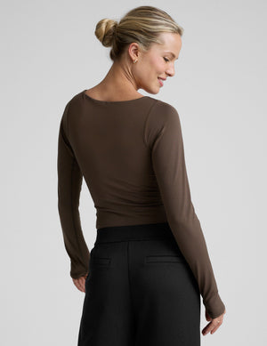 woman wearing a brown long-sleeve boat neck bodysuit and black pants against a plain background. 