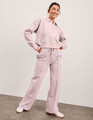 Woman wearing a pink collared pullover and pintuck wide leg sweatpants with white shoes on a plain background. 