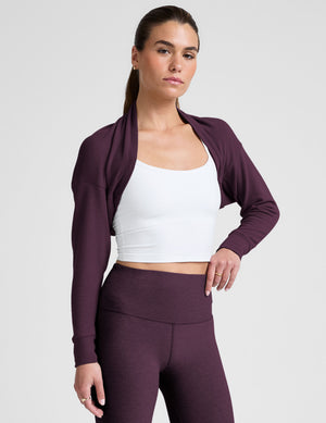 model wearing a white cropped tank and purple leggings with a purple shrug on a light background. 