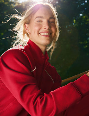 woman wearing a red zip-up jacket with white piping against a nature background. 