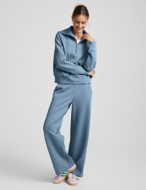 Woman wearing a light blue quarter zip pullover and pants against a plain background. 