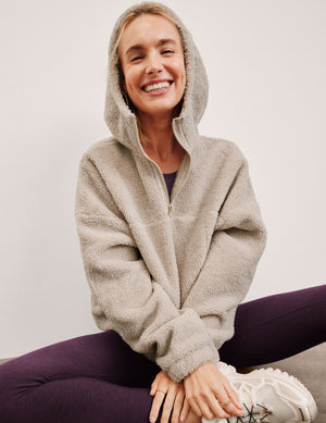 model wearing a beige fleece hoodie sitting against a plain background. 