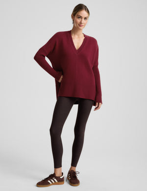 Woman wearing a red v-neck long length sweater and brown leggings against a plain background. 