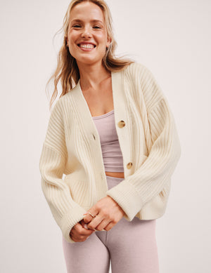 woman wearing a cream cardigan over a pink top and leggings on a plain background. 